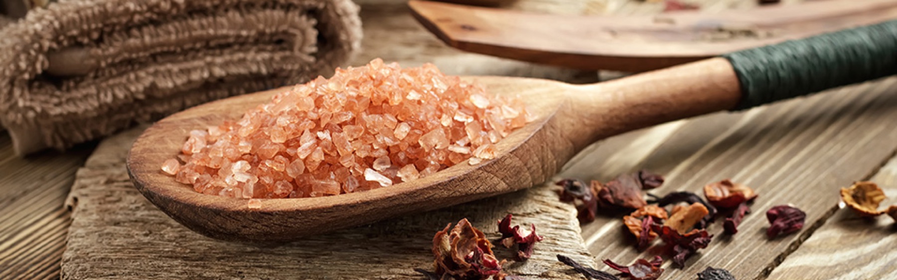 Pink Himalayan salt heaped on a wooden spoon, dried petals scattered on the timber beside it