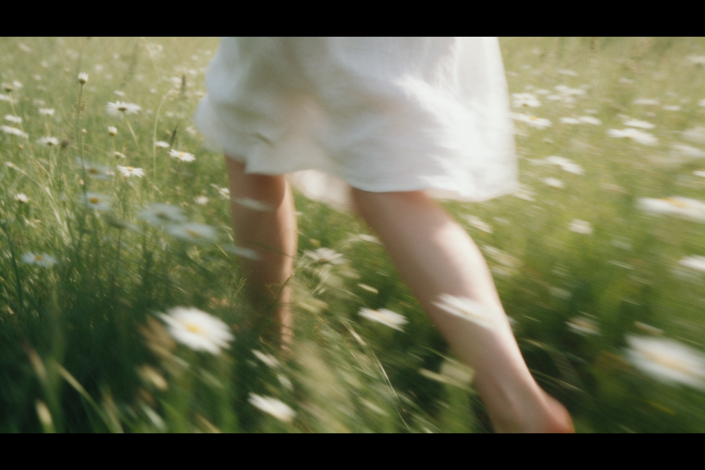 Bare legs and the hem of a linen dress mid-stride through tall grass and daisies